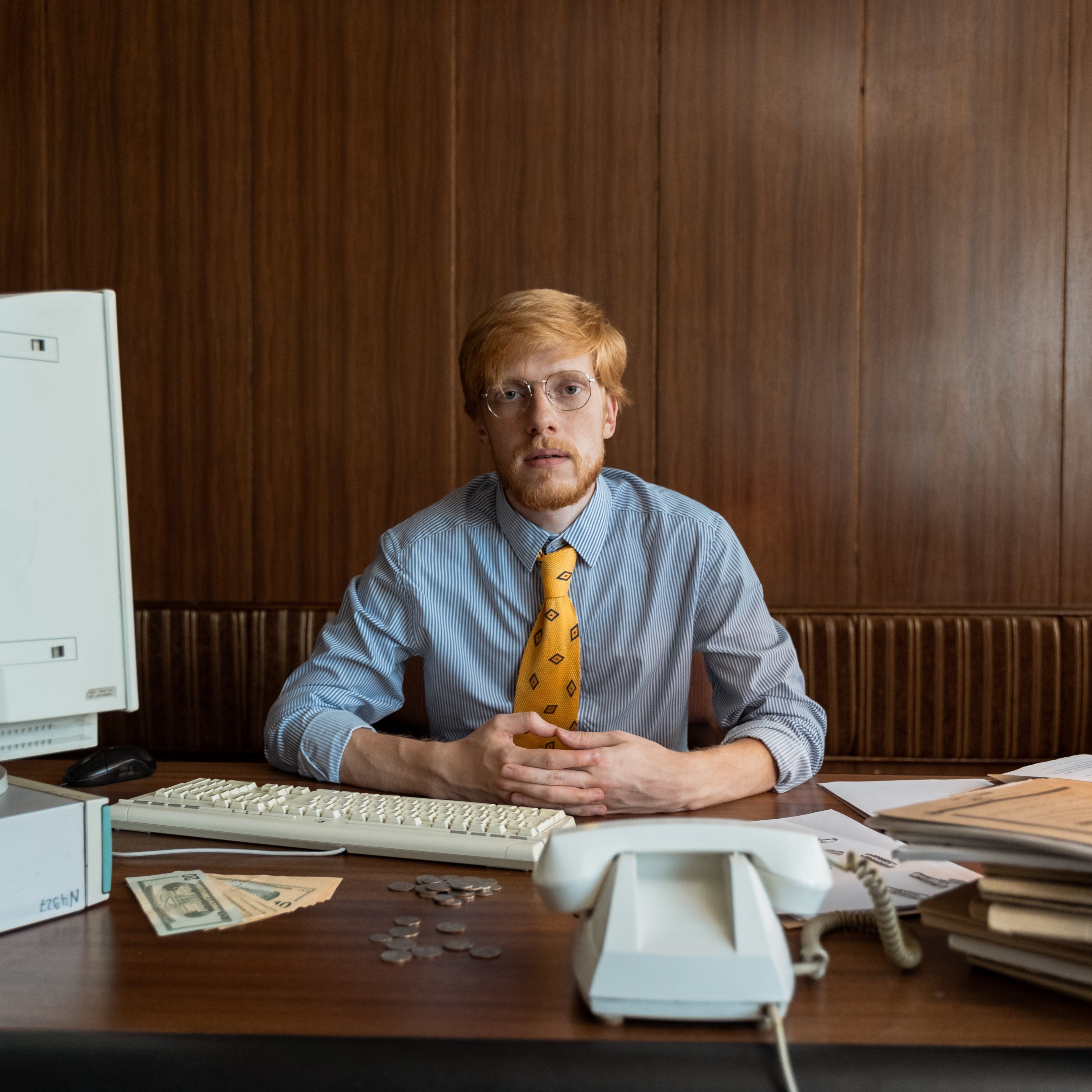 a man with short red hair and glasses stares at the camera judging these choices despite his messy desk.